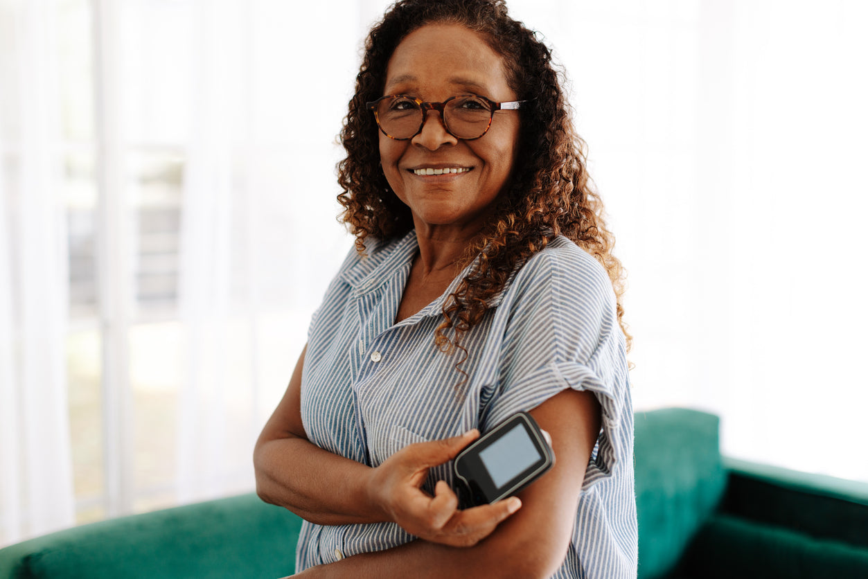 Portrait of a cheerful woman in striped shirt applying a blood sugar monitor on her arm, standing in a bright living room.