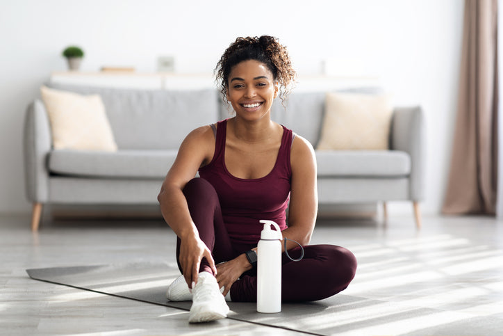 Fit young woman in burgundy activewear resting after exercise at home, sitting on a yoga mat with a water bottle.